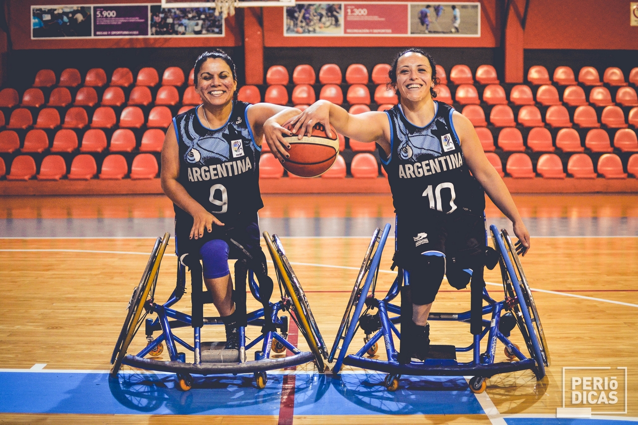 Dos mujeres en silla de rueda, con indumentaria deportiva del seleccionado de básquet argentino, agarrando una pelota de básquet entre las dos mientras sonríen