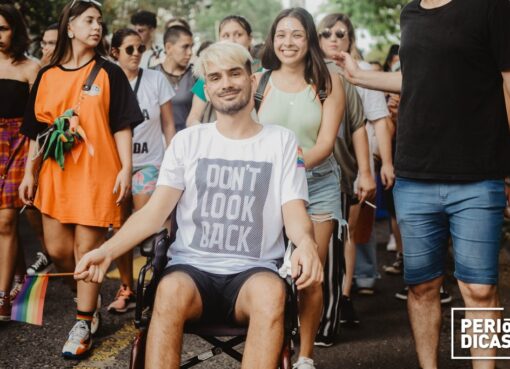 Joven con discapacidad en silla de ruedas, junto a amigues, sosteniendo una pequeña bandera del orgullo, sonriendo, en la Marcha del Orgullo 2021 de Santa Fe.