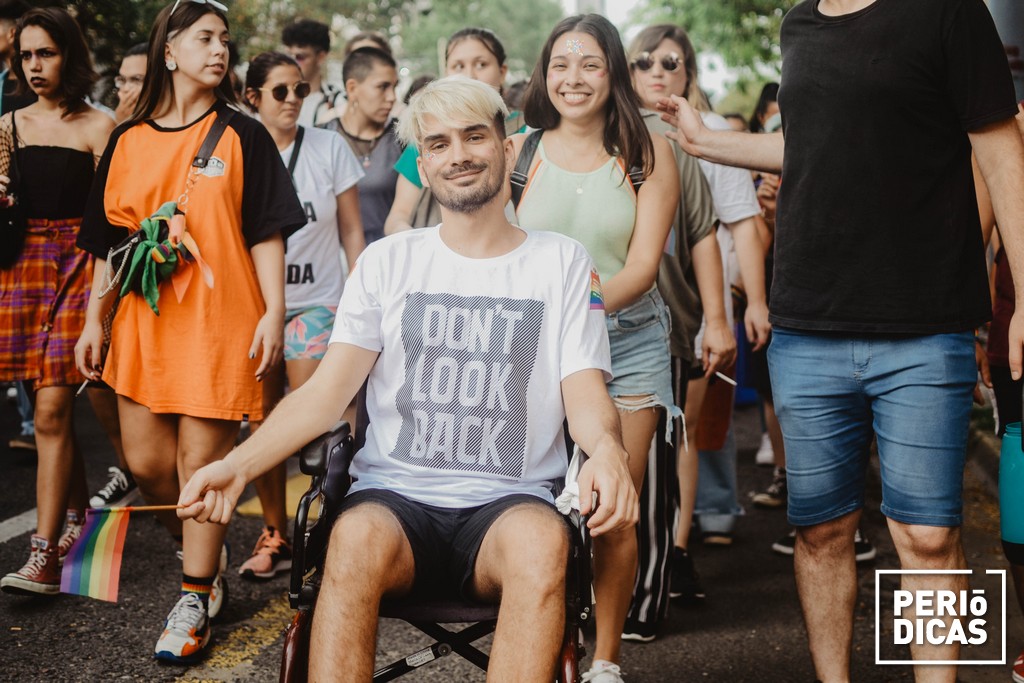 Joven con discapacidad en silla de ruedas, junto a amigues, sosteniendo una pequeña bandera del orgullo, sonriendo, en la Marcha del Orgullo 2021 de Santa Fe.